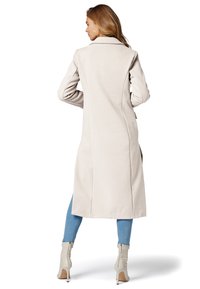 Manteau long et ajusté en tissu beige clair avec une finition texturée. Doté d'un col cranté et de deux poches latérales. Porté sur un jean bleu et des bottes.