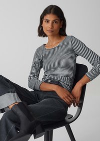 Schwarz-weiß gestreifter Langarm-Oberteil mit rundem Ausschnitt, kombiniert mit hoch taillierten dunklen Jeans und schwarzen Knöchelstiefeln, sitzt auf einem Stuhl.