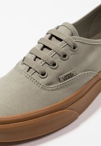 Gros plan d'une sneaker Vans en toile taupe avec des lacets assortis et une semelle en caoutchouc marron sur fond blanc.