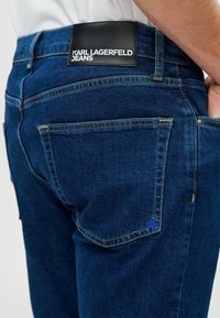 Jeans de ganga azul escuro com um acabamento texturizado. Apresenta um patch de logotipo preto na cintura e costuras amarelas contrastantes nos bolsos.