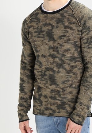 Homme portant un pull à manches longues à motif camouflage en tissu texturé et un jean bleu, debout devant un fond uni.
