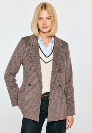 Kobieta z blond bobem, ubrana w brązową kratowaną marynarkę, kremową kamizelkę na sweterku, jasnoniebieską koszulę i ciemne jeansy na białym tle.