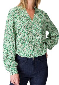 Femme portant un chemisier boutonné à fleurs vert et blanc avec de longues manches bouffantes et un jean bleu foncé.