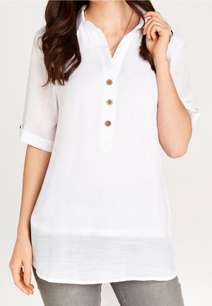 Femme portant un chemisier blanc ample avec trois boutons en bois, manches retroussées, et un pantalon gris, posant avec une main près du visage.