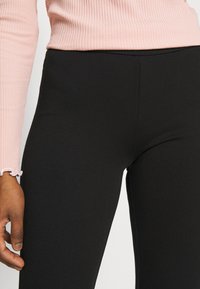 Person wearing a light pink ribbed long-sleeve top with lettuce-edge cuffs and fitted black pants, hand relaxed by side.