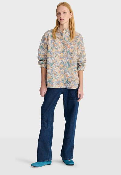 Femme aux longs cheveux blonds portant une chemise ample à fleurs pastel, un jean large bleu foncé et des chaussures plates bleu sarcelle, debout devant un fond uni.