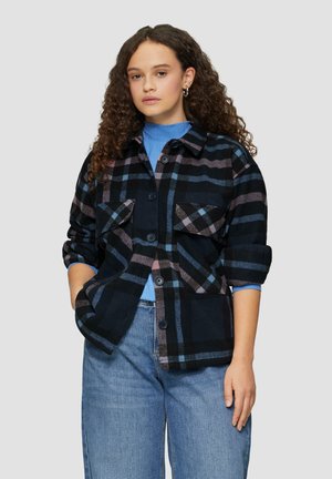 Zwarte en blauwe geruite shirtjas met twee borstzakken, zwarte knopen en opgerolde mouwen, gedragen over een lichtblauw topje en gecombineerd met denim.