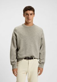 Maglione grigio a maglia con scollatura rotonda, maniche lunghe e polsini a coste, abbinato a pantaloni beige chiaro e una cintura marrone.