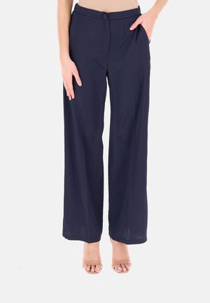Pantaloni a gamba larga blu navy con bottone e cerniera frontale, indossati con tacchi aperti color nude, una mano in tasca, su sfondo semplice.