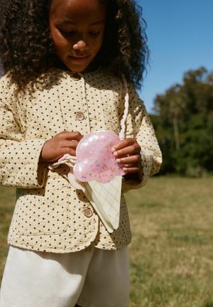 Fetiță tânără în aer liber ținând o poșetă roz în formă de cornet de înghețată, purtând o geacă bej cu buline și pantaloni albi sub un cer albastru senin.