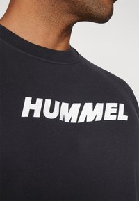 Sort bomulds t-shirt med rund hals, der har et stort hvidt "HUMMEL" logo på tværs af brystet. Glen glatt tekstur med korte ærmer.