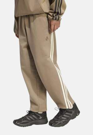 Personne portant un pantalon de survêtement beige Adidas avec des bandes blanches et des baskets Adidas noires, montrée de la taille aux pieds sur un fond uni.
