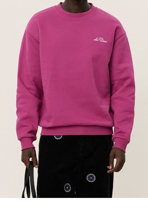 Personne portant un sweat-shirt rose vif avec un petit texte blanc sur la poitrine et un pantalon noir avec des motifs floraux bleus et blancs, tenant une sangle noire.