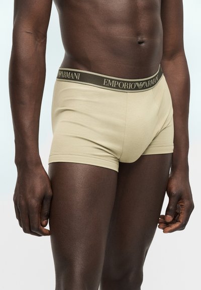 Ανδρικό μοντέλο με μπεζ μποξεράκια Emporio Armani με λογότυπο στη μέση, εμφανιζόμενο από τη μέση έως το επάνω μέρος των μηρών, πάνω σε μονόχρωμο φόντο.