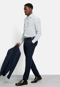 Hellblaues, gemusterte Hemd mit Knöpfen, kombiniert mit dunkelblauer Hose. Dunkelbraune Lederschuhe und ein dunkelblazer, der in der Hand gehalten wird.