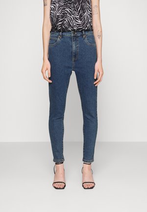 Skinny blue denim jeans with a mid-rise waist, five pockets, and subtle stitching details, paired with a patterned black and white top and black heels.