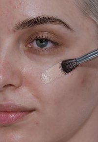 Une fondation crème beige est appliquée sur la peau avec un pinceau, mettant en valeur une texture lisse sur la joue. La peau a un fini naturel et brillant.