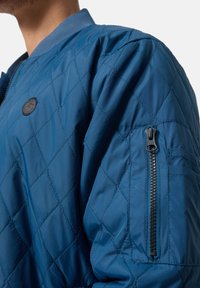 Man draagt een doorgestikte blauwe bomberjack met ruitvormige stiksels, een ritszak op de bovenarm en een ronde zwarte patch op de borst.