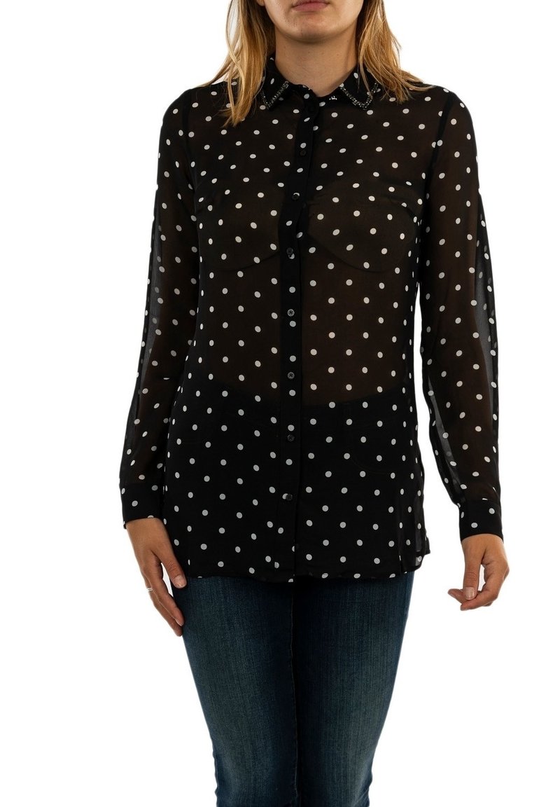 Femme portant un chemisier noir à manches longues, boutonné, à pois blancs et un jean bleu, debout devant un fond blanc.