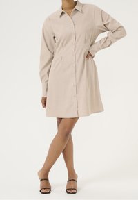 Robe chemise beige à boutons, manches longues, coupe décontractée et col. Texture lisse, longueur genou, associée à des sandales noires à lanières.