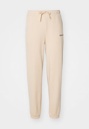 Pantalons de survêtement en polaire beige avec une taille élastique, un cordon de serrage ajustable et des poignets ajustés. Présente un petit logo noir sur le côté.