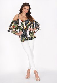 Blomstret off-shoulder bluse med sort baggrund, grønne og orange bladmønstre, parret med hvide skinny jeans og gyldne sandaler med stropper.