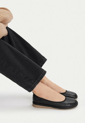 Osoba sedící, oblečená v černých širokých kalhotách a černých kožených slip-on balerínkách s béžovými gumovými podrážkami na bílém pozadí.