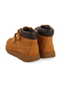 Bottes de cheville en daim marron avec un col en cuir marron, languette rembourrée, sangle à crochet et boucle, et semelle en caoutchouc texturé. Coutures blanches en accents.