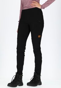 Black, fitted outdoor pants with textured fabric, zippered pockets, and a brown logo patch; paired with black hiking boots.