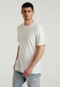 T-shirt a maniche corte di un leggero color beige, realizzato in un morbido tessuto con colletto rotondo, dotato di una piccola applicazione del logo sul lato sinistro del petto e vestibilità comoda.