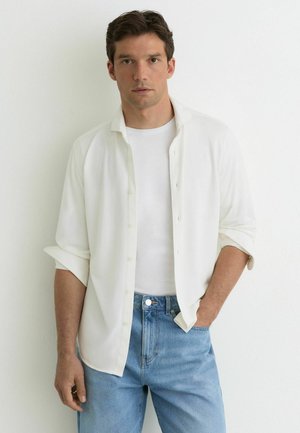 Mladý muž v bílé košili přes bílé tričko a modrých džínách stojí před jednobarevnou bílou zdí.