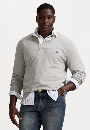 Man in een gelaagde lichtgrijze rugbyshirt over een blauw-wit gestreept overhemd met knopen, blauwe jeans en een bruine leren horloge met een donker gezicht.