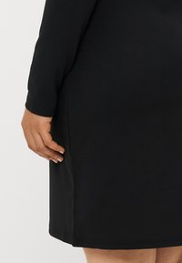 Μαύρο ελαστικό φόρεμα με μακριά μανίκια και εφαρμοστή σιλουέτα, που διαθέτει καθαρές γραμμές και ομαλή υφή. Ελαφρά ορατή λεπτομέρεια στη ραφή πλε�υράς.