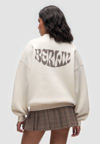 Felpa oversize color crema con "BERLIN" in lettere marroni ondulate sul retro, abbinata a una gonna a pieghe in tartan marrone.