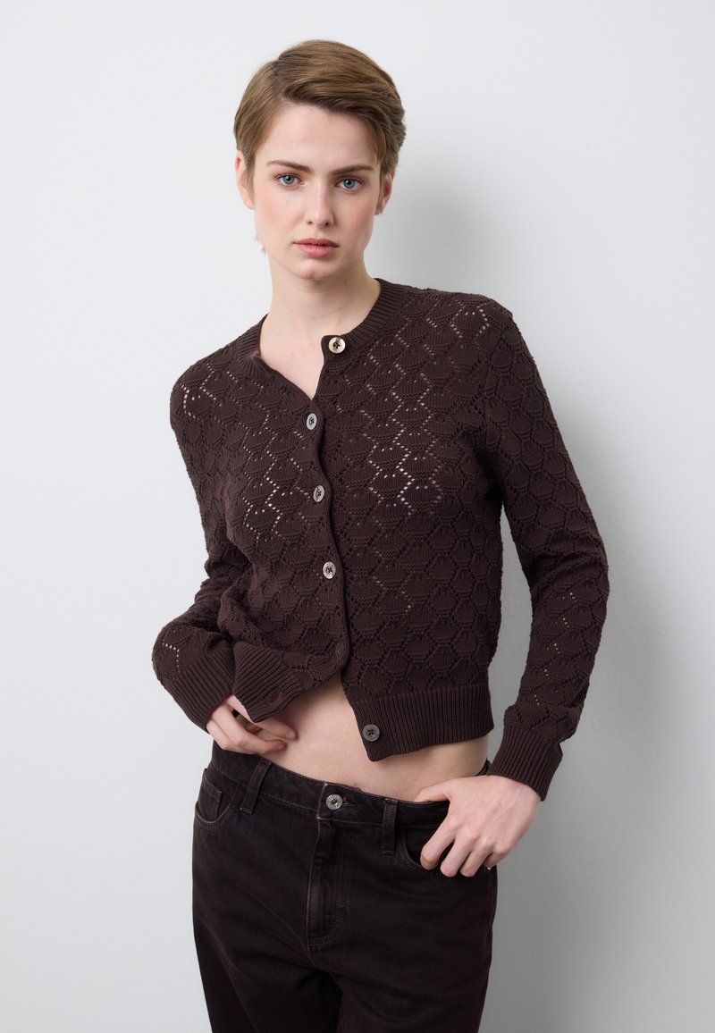 Braune, geometrisch gemusterte Strickjacke mit Rundhalsausschnitt, Knopfleiste vorne und gerippten Bündchen. Kombiniert mit dunkler Hose, die einen offenen Bauchbereich zeigt.