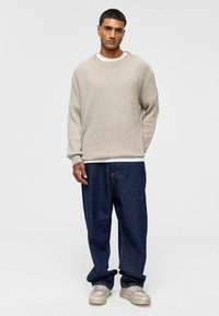 Beige gebreide sweater met een geribbelde textuur, gecombineerd met losse donkerblauwe spijkerbroeken. Model draagt witte sneakers en staat tegen een lichte achtergrond.