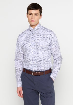 Witte overhemdbloes met een blauw en paarse stippenpatroon, lange mouwen en een puntige kraag. Gecombineerd met donkere broek en een bruine riem.