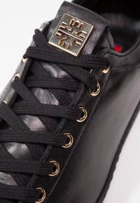 Baskets en cuir noir avec lacets noirs, œillets dorés et un emblème carré doré sur la languette présentant un design symétrique stylisé.