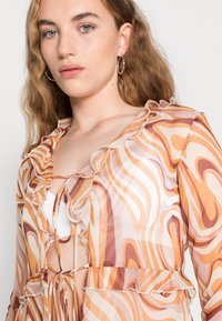 Blusa transparente e estampada com desenhos ondulados em laranja e castanho, bordas franzidas e fecho por laço no decote.