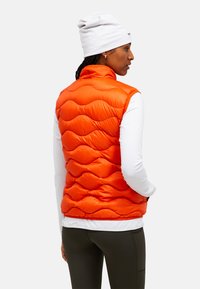 Oranje, gewatteerde puffer vest met een hoge kraag en mouwloos ontwerp. Draag het over een wit longsleeve shirt, dat een gladde textuur heeft.