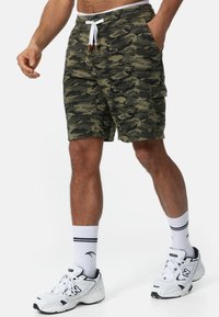 Camo-shorts in groen en zwart met een tailleband met trekkoord, zijzakken en een cargozak aan de rechterzijde. Gecombineerd met witte sneakers.