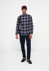 Camicia a quadri blu con accenti bianchi e rossi, abbinata a pantaloni scuri e mocassini neri. Accessorio: cappellino bordeaux e borsa a tracolla.