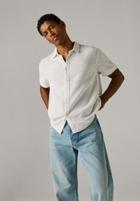 Άνδρας με κοντά σγουρά μαλλιά, φοράει άσπρο πουκάμισο με κοντά μανίκια και κουμπιά και ανοιχτό μπλε τζιν, στέκεται με τα χέρια πίσω από την πλάτη.