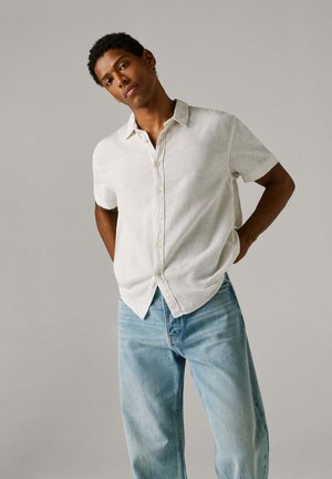 Mies, jolla on lyhyet kiharat hiukset, pukeutunut valkoiseen lyhythihaiseen nappipaitaan ja vaaleansinisiin farkkuihin, seisoo käsivarret selän takana.