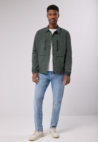 Olivově zelená lehká bunda se zipovými kapsami na hrudi, spárovaná s světle modrými džíny a krémovými teniskami. Hladká textura, knoflíkové zapínání.