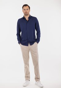 Marineblå langermet skjorte med knapper foran, kombinert med lysebeige linbukser og hvite sneakers, vist mot en hvit bakgrunn.