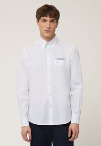 Chemise blanche à boutons avec un col pointu, dotée d'une poche poitrine à motif et de manches longues. Tissu lisse, coupe classique, design minimaliste.