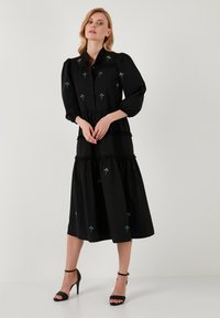 Rochie neagră cu mâneci bufante lungi și nasturi pe față, având broderie florală albastră și detalii cu volane stratificate. Țesătură netedă, lungime până la gleznă.