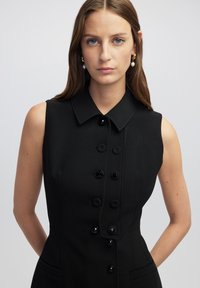 Schwarzes ärmelloses Kleid mit einem strukturierten Kragen, ausgestattet mit sechs großen Knöpfen, figurbetont, glatte Textur und zwei seitlichen Taschen.
