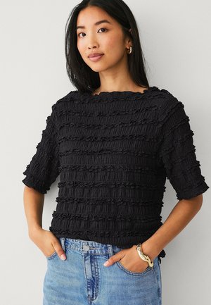 Vrouw die een gestructureerd zwart topje met korte mouwen en horizontale ruches draagt, lichtblauwe jeans, een gouden armband en gouden creolen, staand met handen in de zakken.